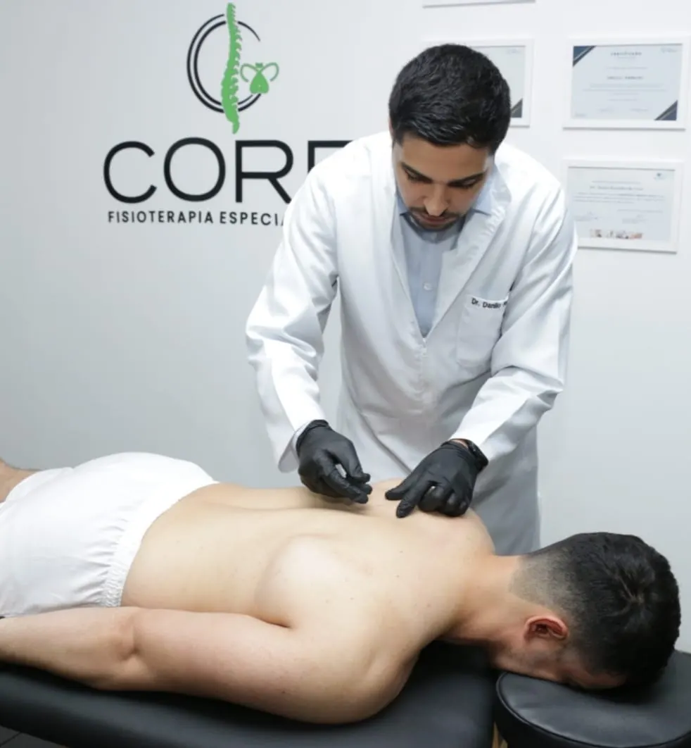 Fisioterapeuta de jaleco branco atendendo um paciente deitado de bruços na maca, com o logotipo da Core na parede ao fundo