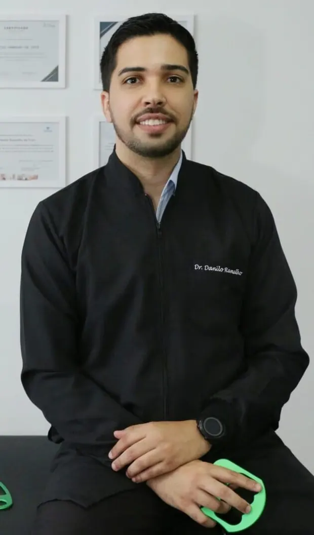 Dr. Danilo Ramalho, fisioterapeuta da Core, de jaleco preto, diante da parede de certificados da clínica