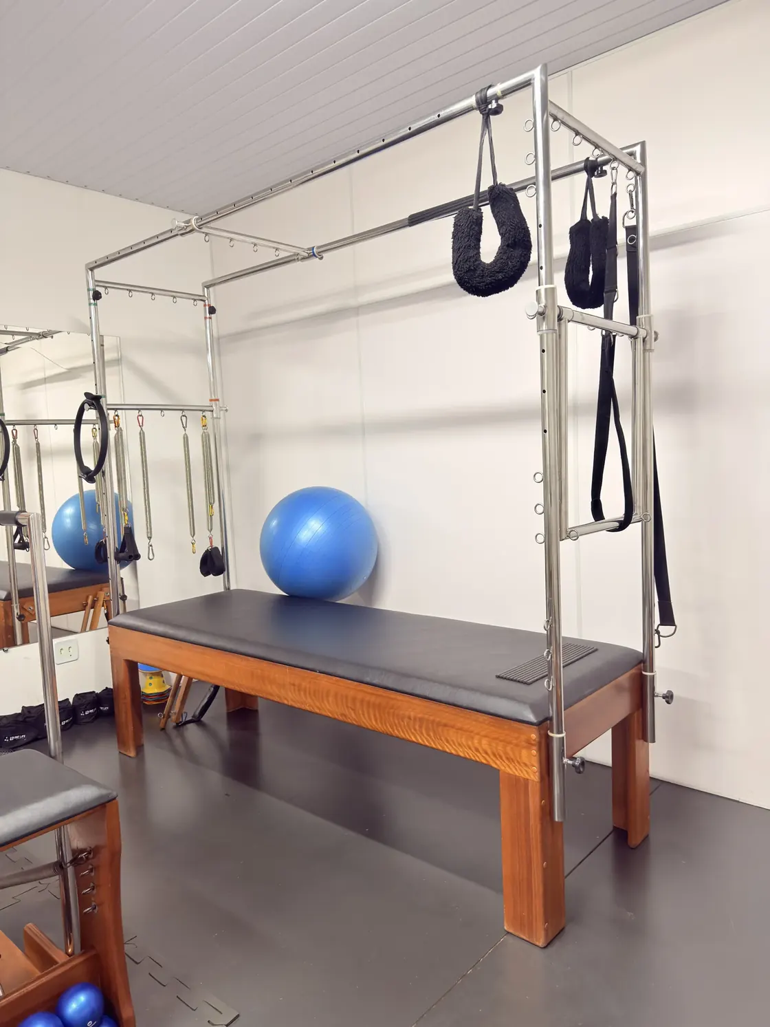 Cadillac de pilates com maca preta, molas e alças suspensas; ao fundo, bola suíça azul e outro aparelho