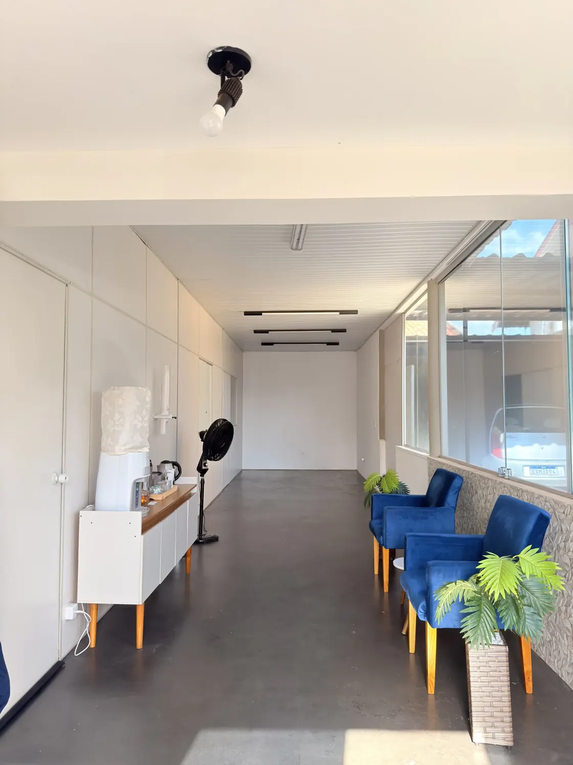 Corredor da clínica com poltronas azuis, vasos de planta e bebedouro junto à parede branca