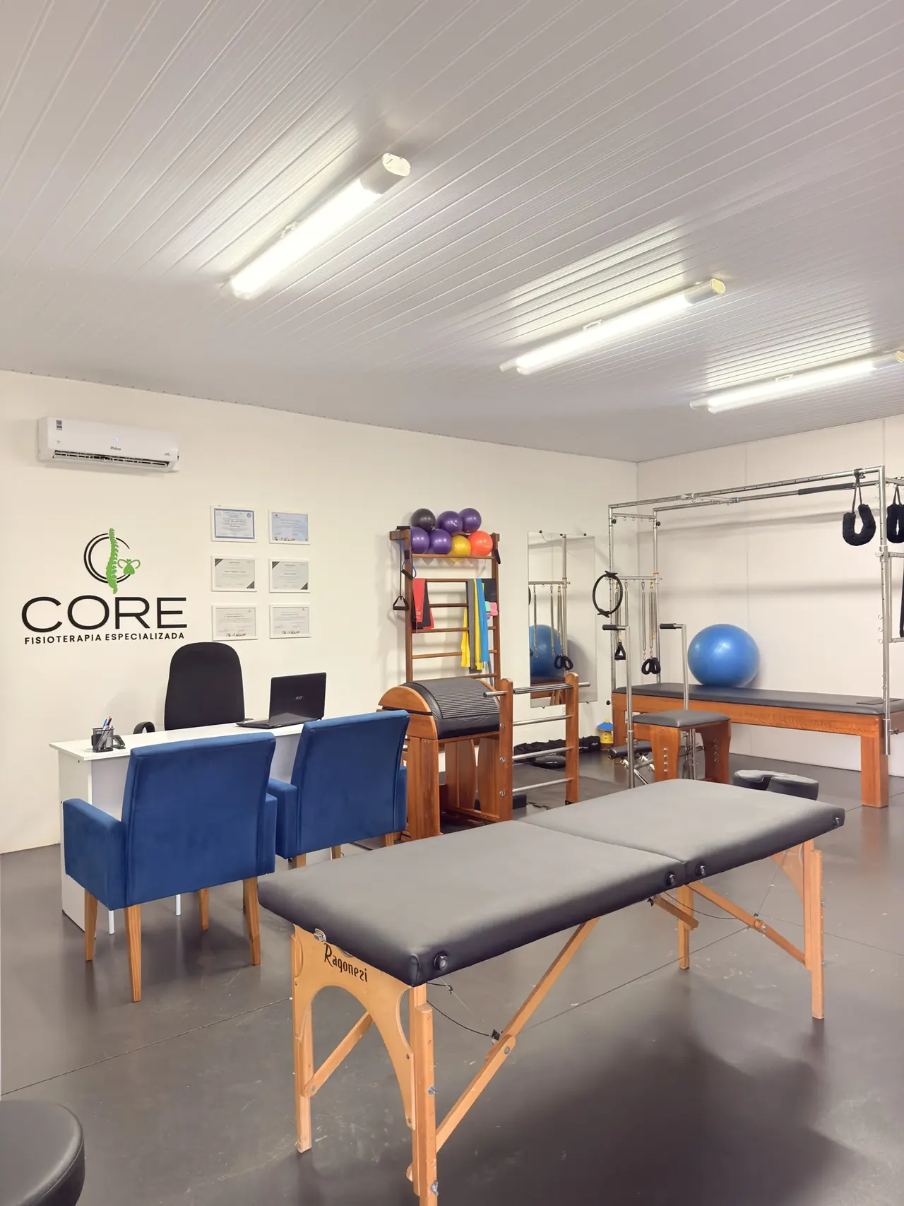 Vista geral da clínica: logotipo da Core na parede, poltronas azuis de espera, maca de atendimento e aparelhos de pilates ao fundo