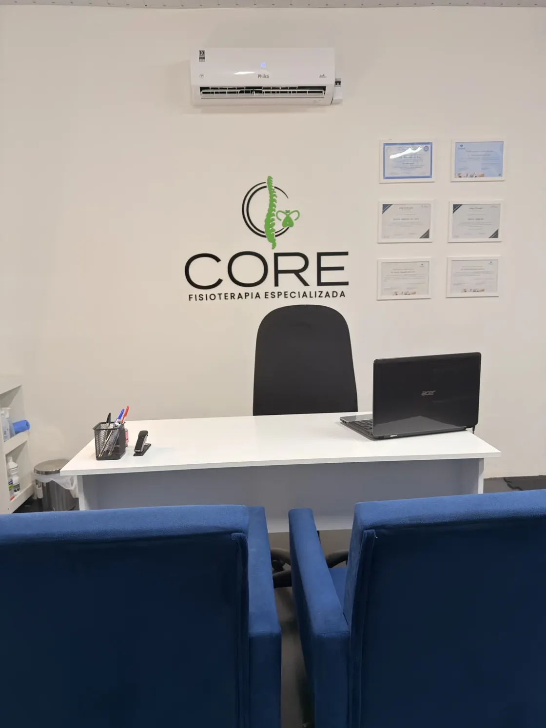 Recepção da Core: mesa branca com notebook, o logotipo da clínica aplicado na parede e certificados emoldurados ao lado