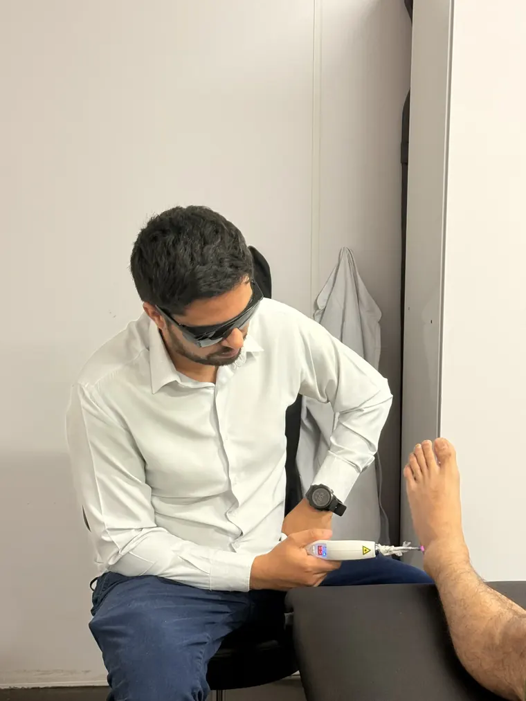 Fisioterapeuta de óculos de proteção escuros aplicando laserterapia no pé de um paciente sentado